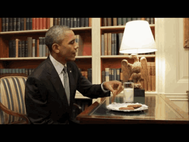 a man in a suit sits at a table with a plate of cookies and a glass of milk