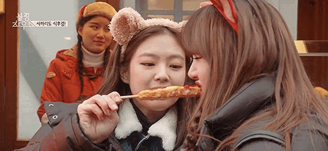 a girl wearing a headband with ears is feeding another girl a stick of food .