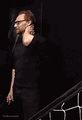 a man wearing glasses and a black shirt is standing with his hands on his hips in a dark room .