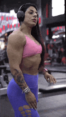 a very muscular woman wearing headphones and a pink top is standing in a gym .