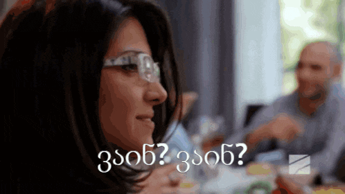 a woman wearing glasses is sitting at a table with a man in the background and the words 3s06 written in white