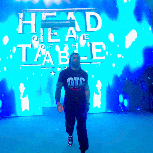 a man wearing a gtc shirt walks in front of a blue screen that says head of the table