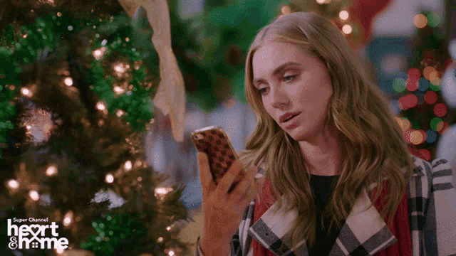 a woman is looking at her phone in front of a christmas tree with the words super channel heart home written on the bottom