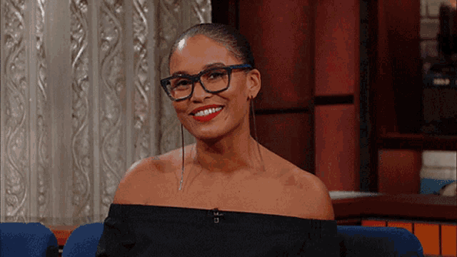 a woman wearing glasses and red lipstick smiles while sitting on a couch