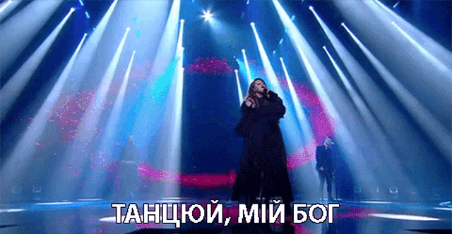 a woman singing into a microphone on a stage with the words " танцуй мій бог " above her