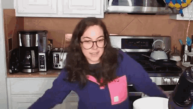 a woman wearing glasses is standing in front of a stove in a kitchen with her arms outstretched .