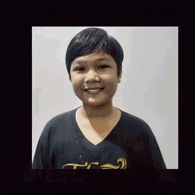 a young boy wearing a black t-shirt is smiling for the camera while standing in front of a white wall .