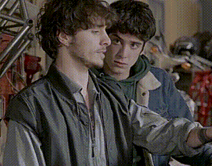 two young men are standing next to each other in a room . one of the men is wearing a green hoodie .