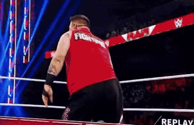 a wrestler in a red tank top is standing in a wrestling ring .
