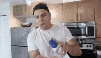 a man in a white t-shirt is holding a blue bottle in his hand in a kitchen .