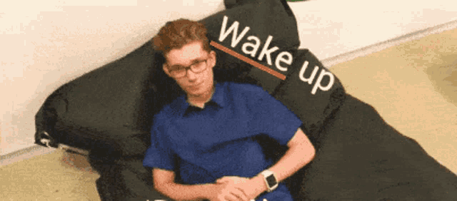 a man in a blue shirt is laying on a black bean bag chair with the words wake up written on it