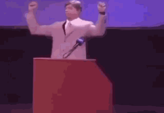 a man in a white suit and tie is standing at a podium with a microphone .