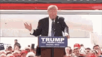 donald trump is giving a speech at a podium while holding a trump sign .