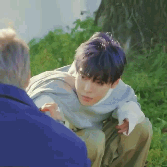 a young man with purple hair is kneeling down in the grass talking to a man .