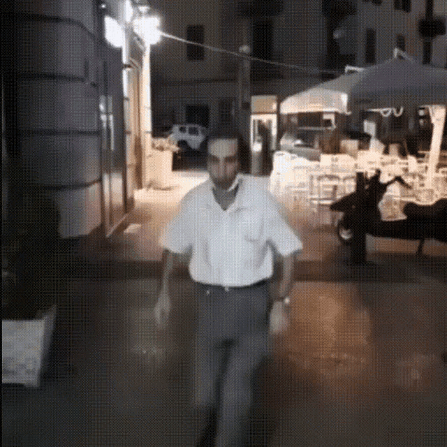 a man in a white shirt and gray pants is walking down a street at night