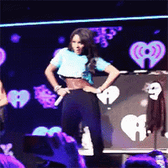 a woman in a blue shirt is dancing on a stage in front of a sign that says iheartradio