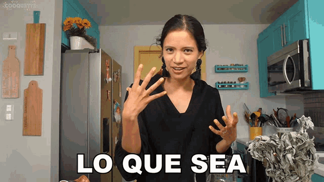 a woman in a kitchen says lo que sea with her hands in the air