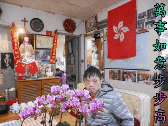 a man sits in a living room with flowers and a clock on the wall