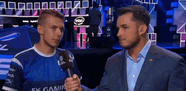 a man in a suit talks into a microphone while another man looks on