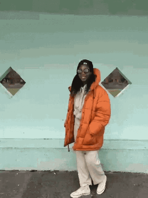 a woman wearing an orange jacket and sunglasses is standing in front of a blue wall .