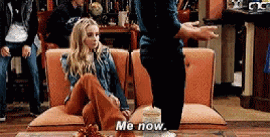 a woman is sitting on a couch with her legs crossed and a man is standing next to her saying me now .