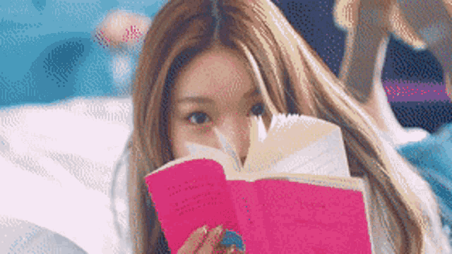 a woman is covering her face with a pink book while laying on a bed .