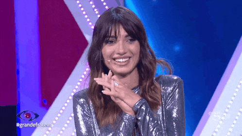 a woman in a sequined top is smiling with the hashtag grandefratello on the screen behind her