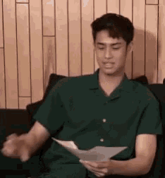 a young man in a green shirt is sitting on a couch holding a piece of paper .