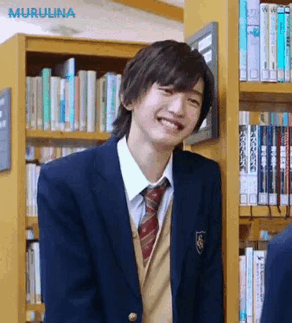 a young man in a school uniform is smiling in front of a bookshelf in a library .