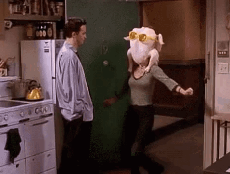 a woman with a turkey on her head is dancing in a kitchen with a man standing behind her .
