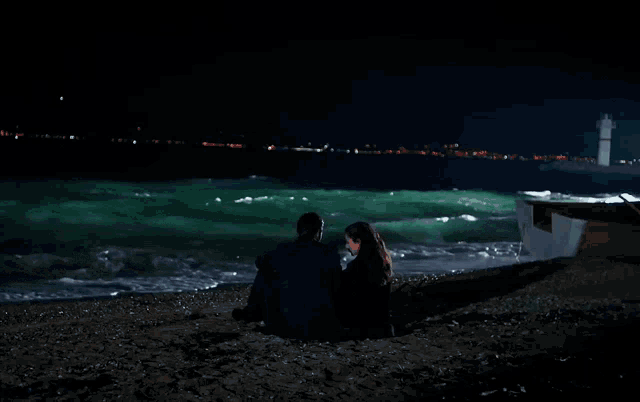 a man and a woman sit on the beach at night looking at the ocean