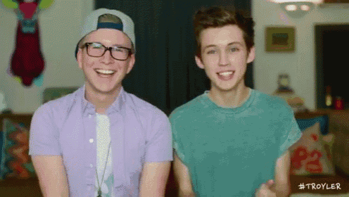two young men are sitting next to each other in a living room and one of them is wearing glasses and a baseball cap ..