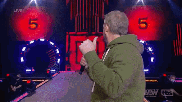 a man in a green hoodie is holding a microphone and giving a thumbs up .