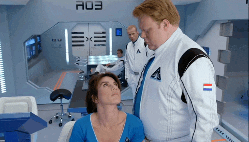 a man in a space suit stands next to a woman in a room with a03 on the wall