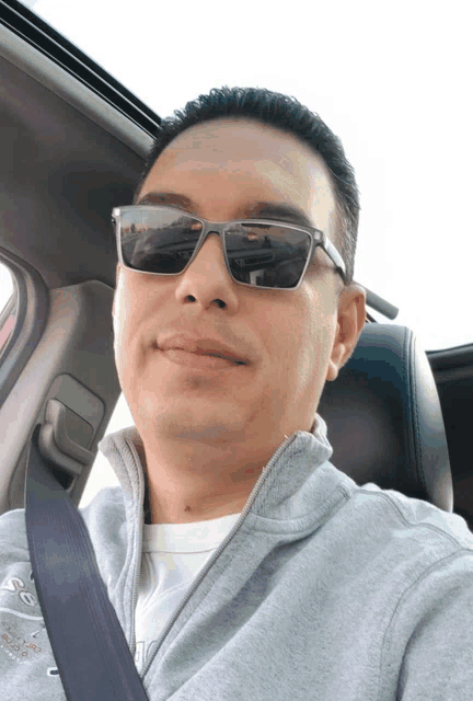 a man wearing sunglasses is sitting in a car with a seat belt around his neck