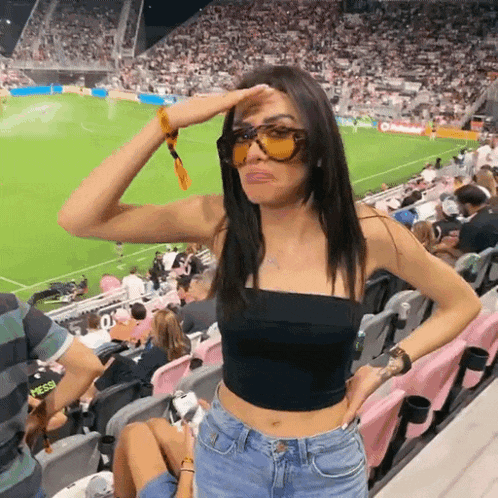 a woman wearing sunglasses and a black top stands in the stands at a soccer game
