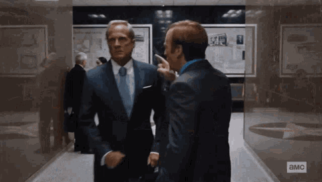 two men in suits and ties are standing in a hallway with a sign that says ' a.m.c ' on it