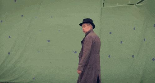 a man wearing a hat and trench coat stands in front of a green screen