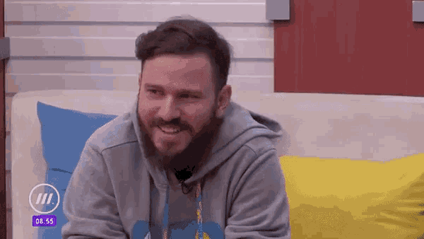 a man with a beard is sitting on a couch with the time of 8:55 on the screen