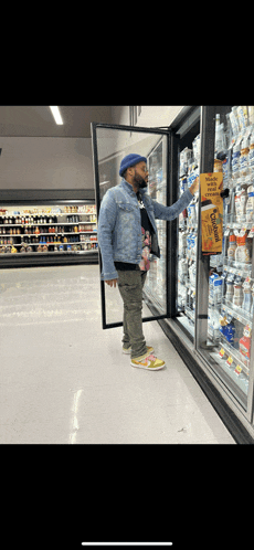 a man in a denim jacket is standing in a grocery store reaching into a cooler for a bag of doritos