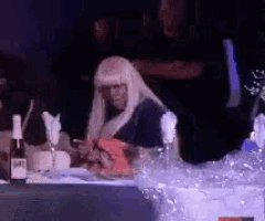 a woman in a blonde wig sits at a table with a bottle of wine