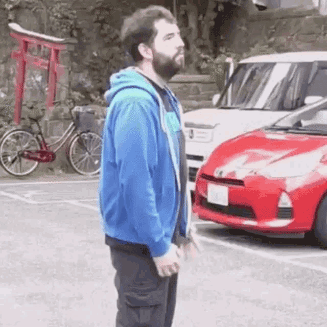 a man in a blue hoodie is standing in a parking lot next to a red car and a bicycle .