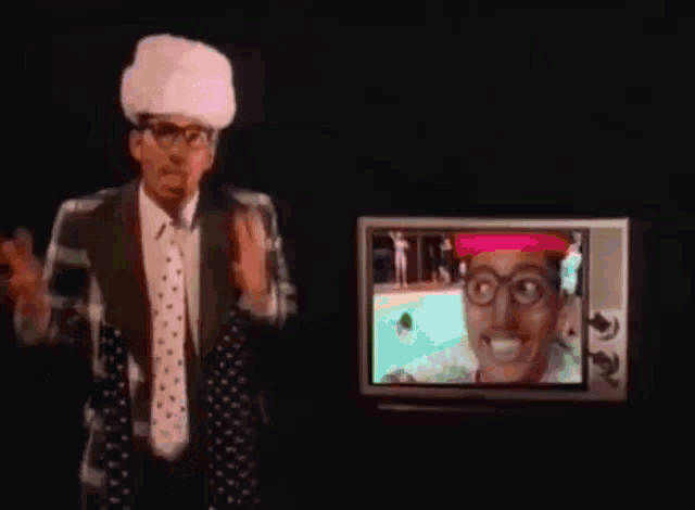 a man with glasses and a pink hat is standing in front of a tv screen