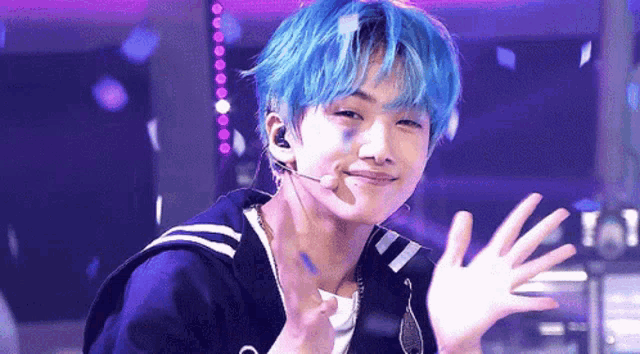 a young man with blue hair is waving at the camera while wearing a microphone .