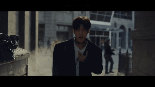 a man in a suit and choker stands in a dark alleyway with a trash can that says ' trash ' on it