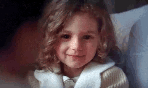 a little girl with curly hair is smiling at the camera while sitting on a bed .
