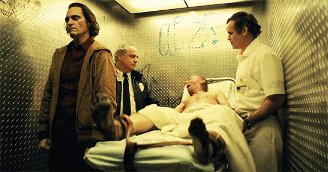 a man in a hospital bed is surrounded by two men and the word us is written on the wall behind him