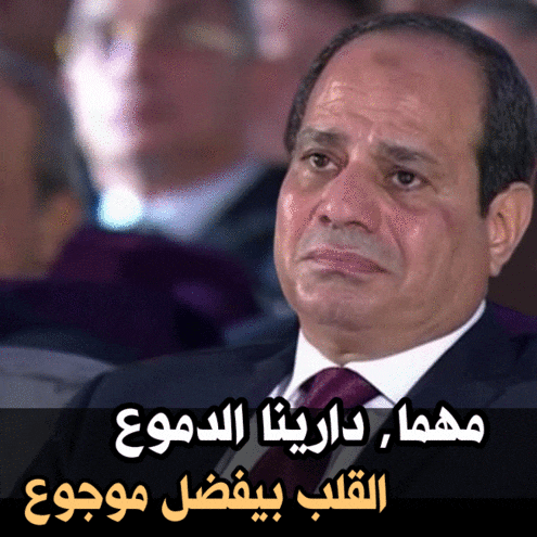 a man in a suit and tie is sitting in front of a sign that has arabic writing on it