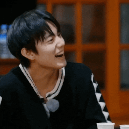 a young man is laughing while sitting at a table with two cups of coffee .
