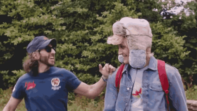 two men are standing next to each other in a field and one of them is wearing a furry hat .